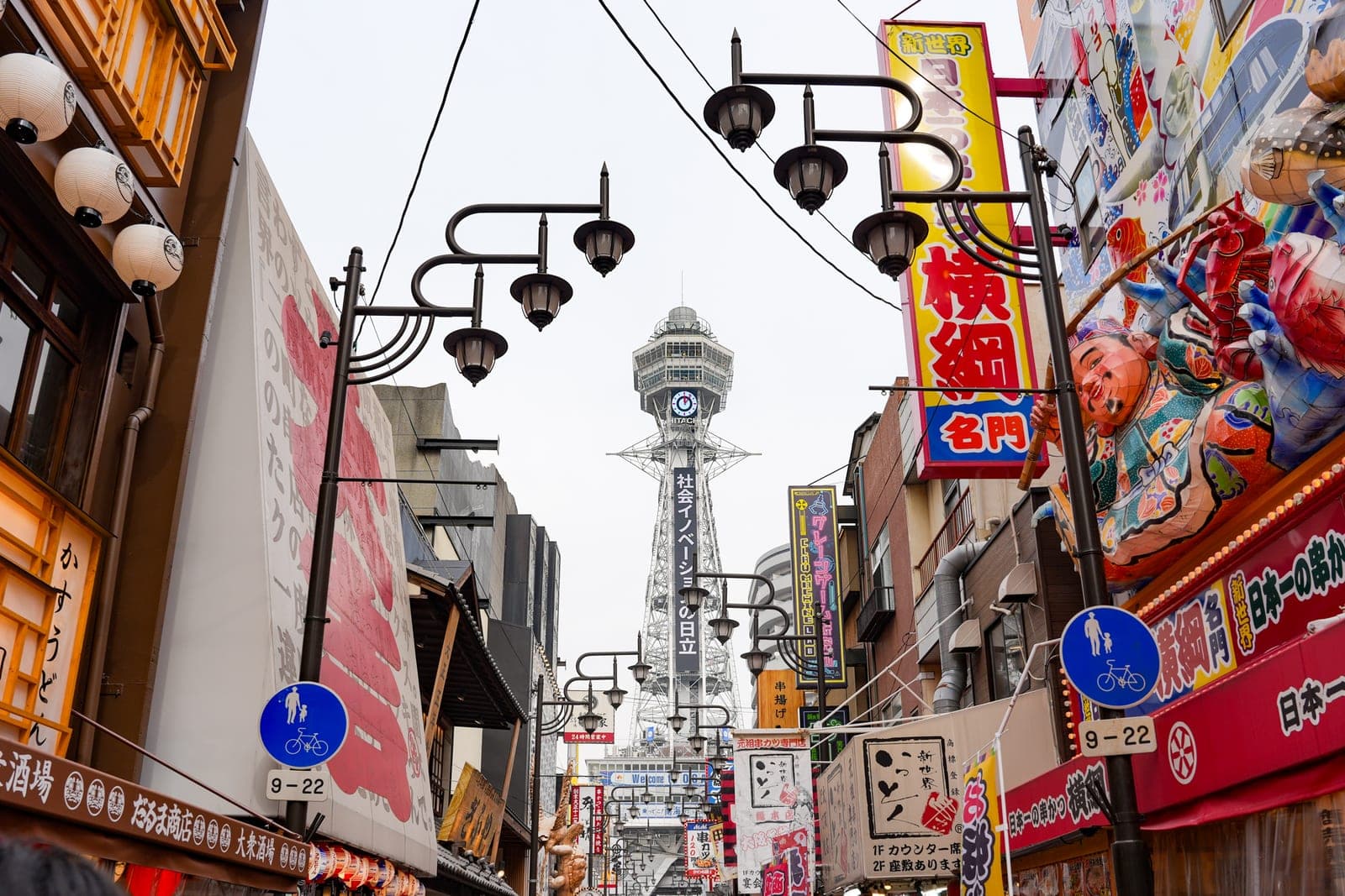 Tsutenkaku tower seen from a Shinsekai street, framed by gas-lamp-style streetlights and dense restaurant signage on both sides